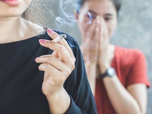Woman in background covers her mouth and nose while woman in the foreground smokes